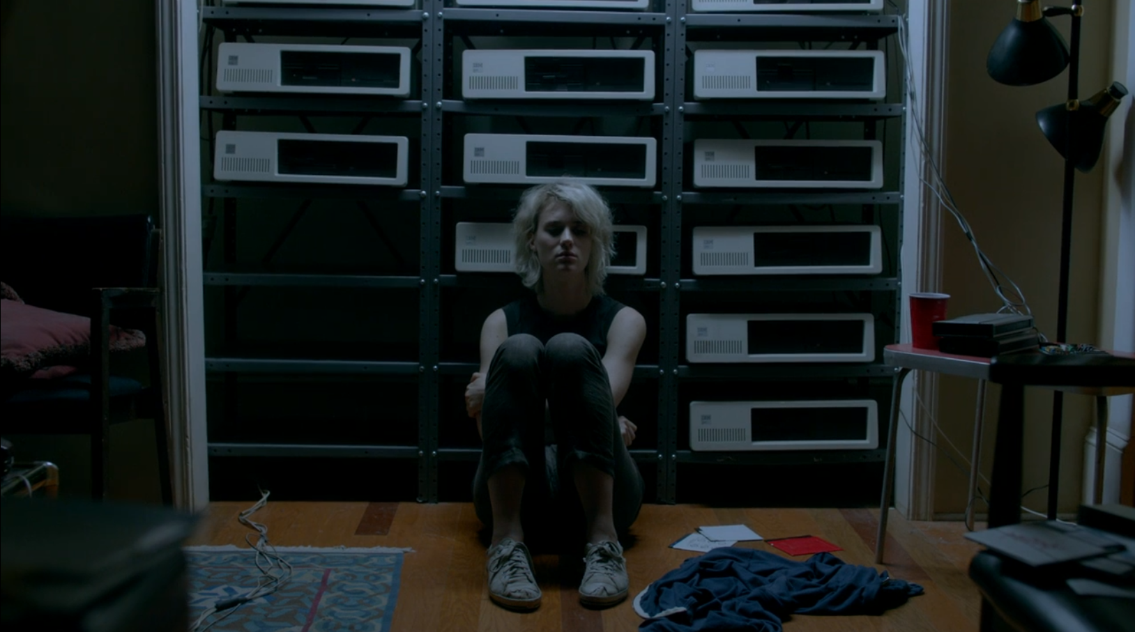 A young woman with dyed blonde hair sits on the floor in front of a wall full of IBM XT servers