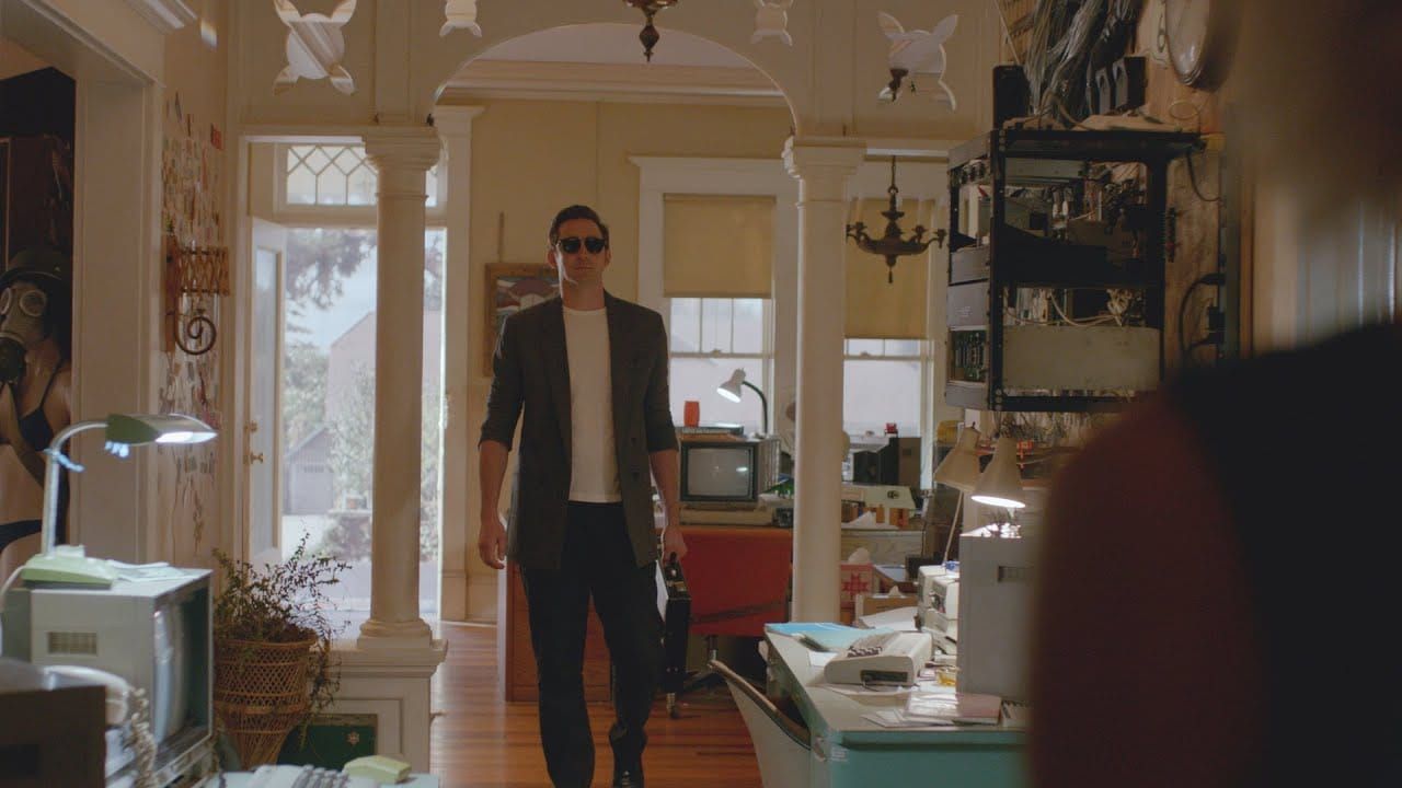 A tall man in dark pants, a gray blazer, a white t-shirt, and sunglasses carries a briefcase into a house full of computers.