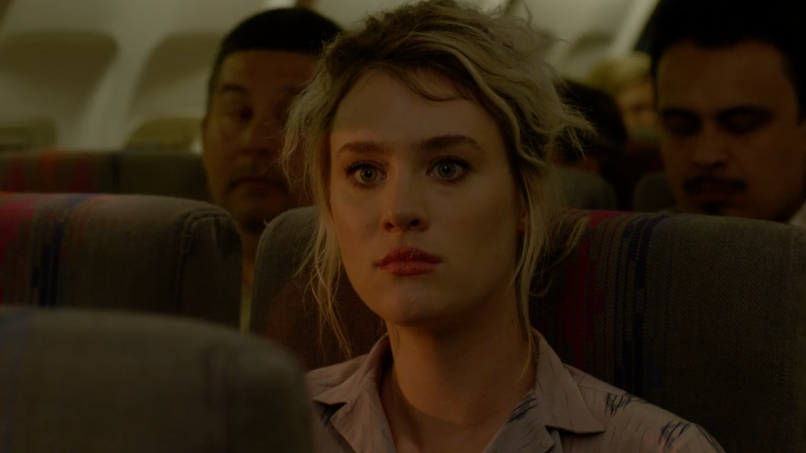 A woman with dyed blonde hair looks expectantly towards the front of the plane for someone to show up.
