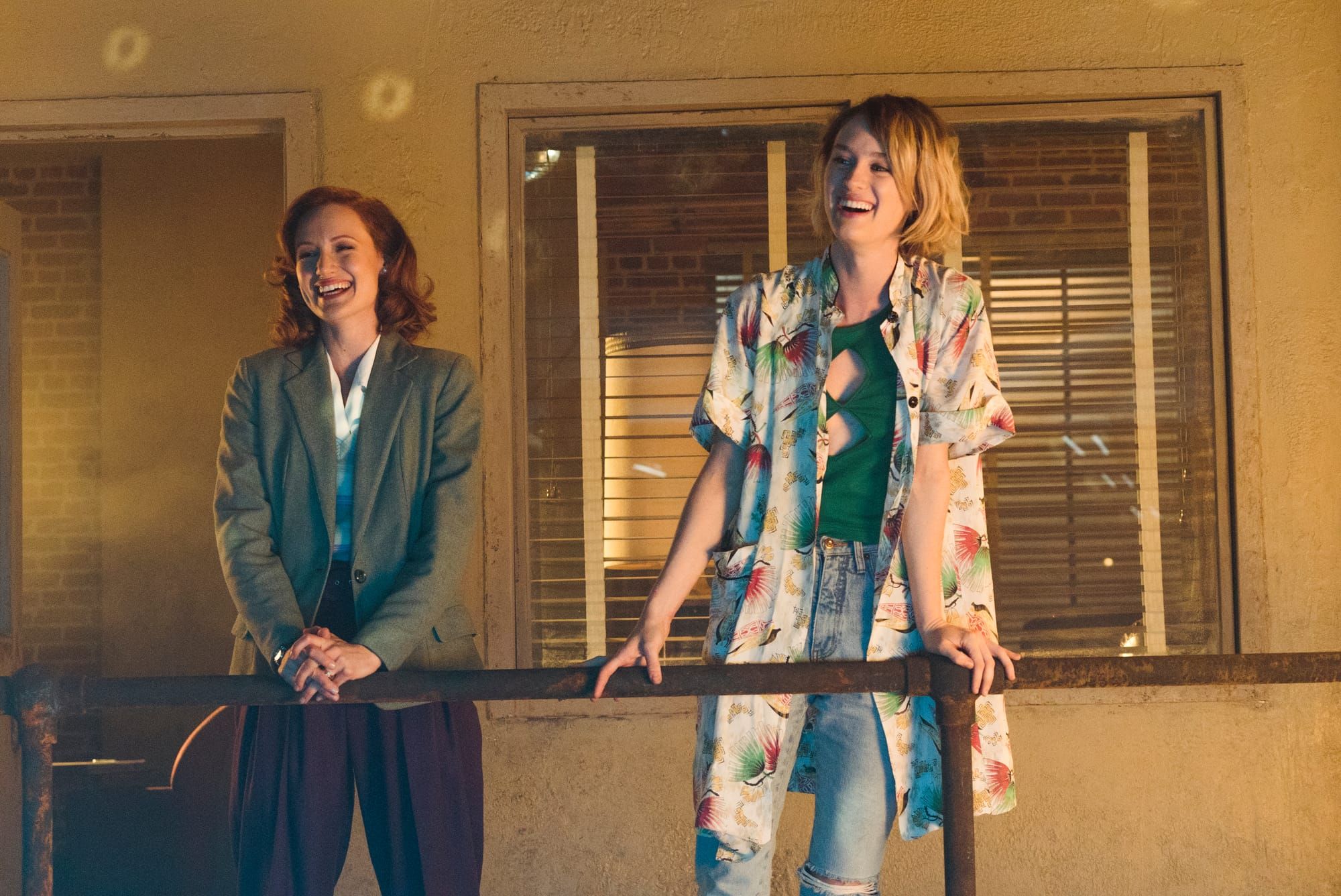 A woman in a long floral print shirt over jeans and green top stands next to a woman in a blazer and dark red pants. They are laughing along with people offscreen.