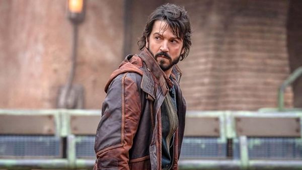A man brown hair and a beard is standing in the road in a town. He is dressed in a brown jacket and looks over his shoulder.