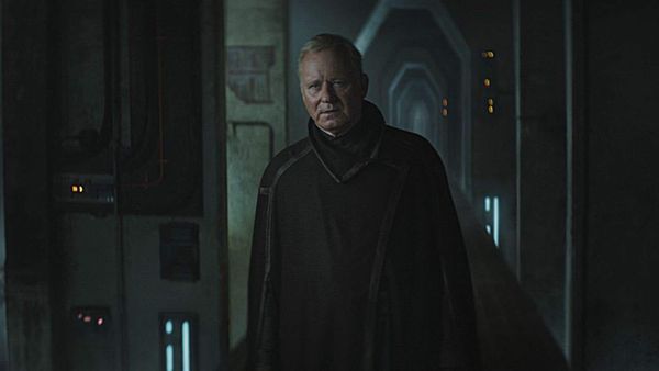 An older man in a dark cloak stands atop a dimly lit narrow walkway without any safety rails.