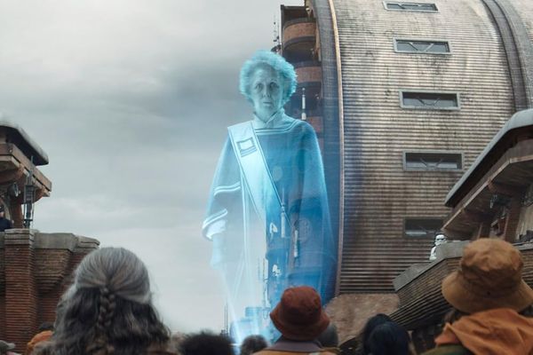 A holographic projection of an elderly woman towers over a group of people in red and orange clothing in an industrial looking city.