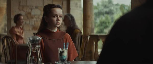 A young girl with brown hair sits in a scenic open air cafe. She wears an orange and red outfit and listens to someone in the foreground. 