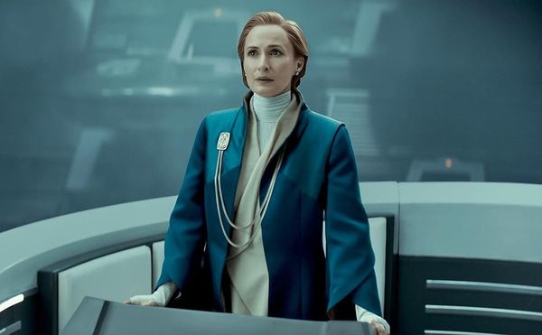 A middle aged woman with red hair stands at a podium in the middle of a Senate chamber and gives a speech. She wears a blue coat over a tan robe.