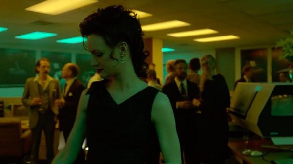 A woman in a stylish black dress with her hair up stands in the middle of party bathed in the worst green light you've ever seen.