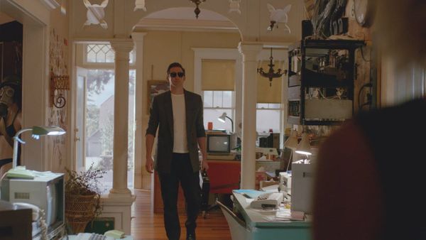 A tall man in dark pants, a gray blazer, a white t-shirt, and sunglasses carries a briefcase into a house full of computers.