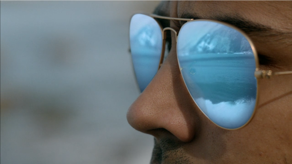 A closeup of a man's face. He's wearing sunglasses, with the reflection of water and a crashing wave shown in the lenses.