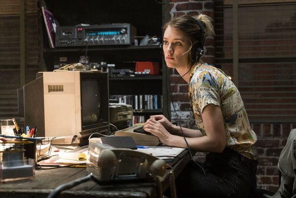 A woman in a colorful shirt with headphones on sits at a computer and looks annoyed at someone offscreen.