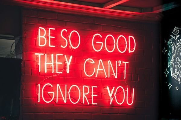 Red neon sign on brick wall reading 'BE SO GOOD THEY CAN'T IGNORE YOU