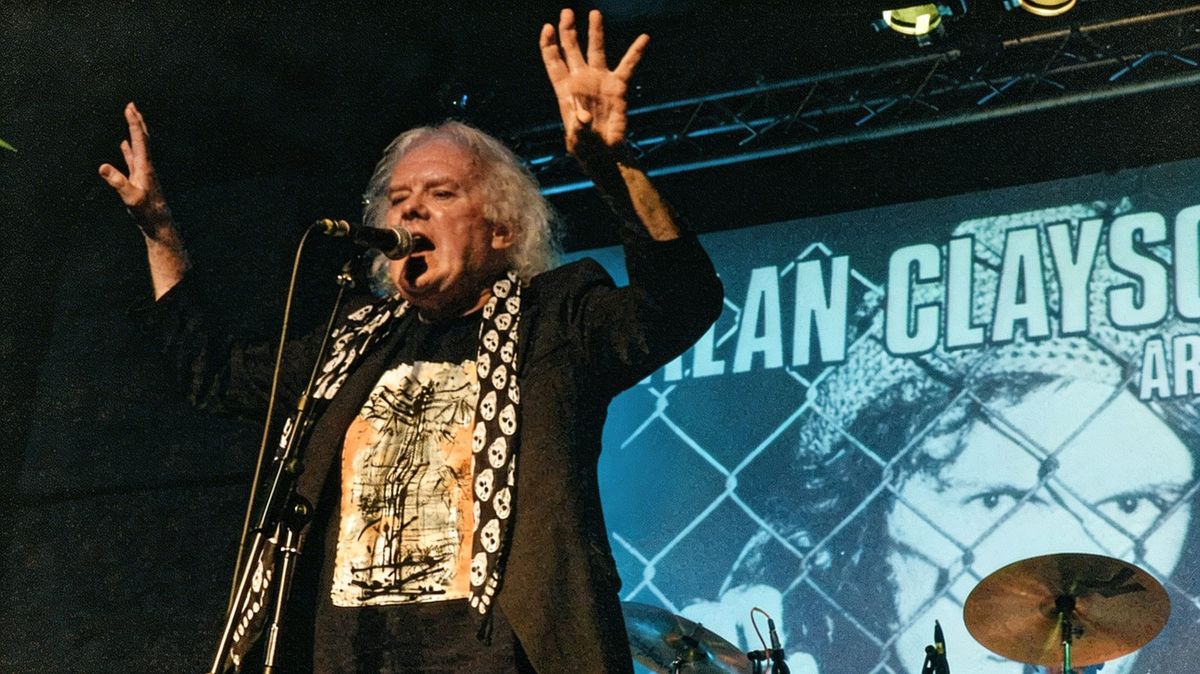 Alan Clayson singing with arms raised at a live concert with Clayson and the Argonauts, with drums and stage banner visible