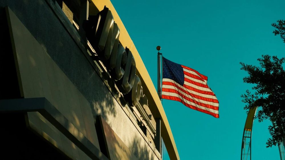 Drapeau américain flottant devant un bâtiment avec le logo 'NUCOR', symbole de puissance économique.