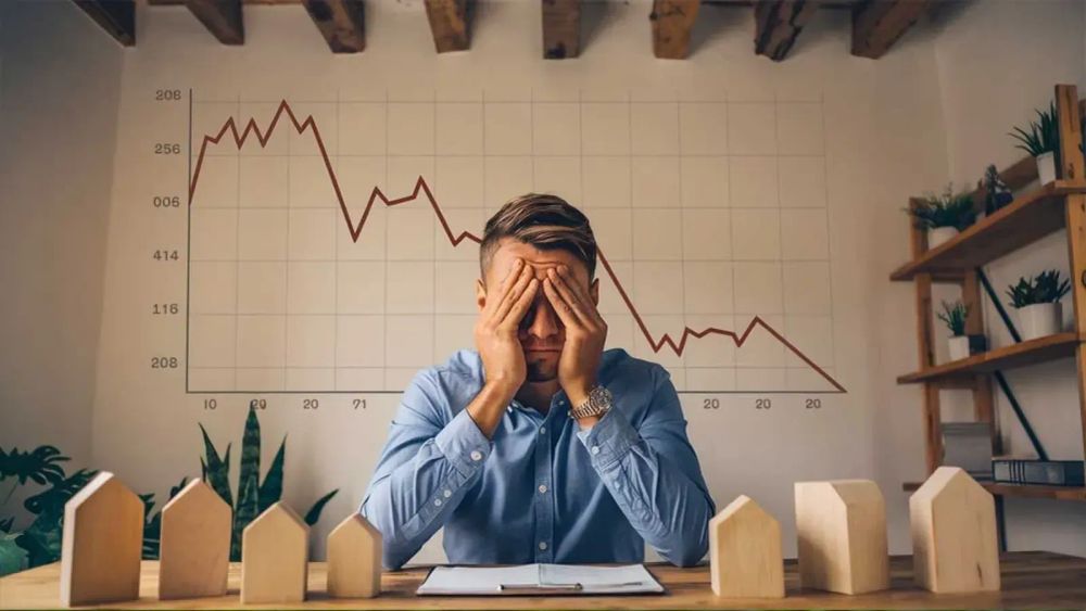 Un homme d'affaires, visiblement stressé, se tient la tête devant un graphique boursier en chute libre et des maquettes de maisons en bois.