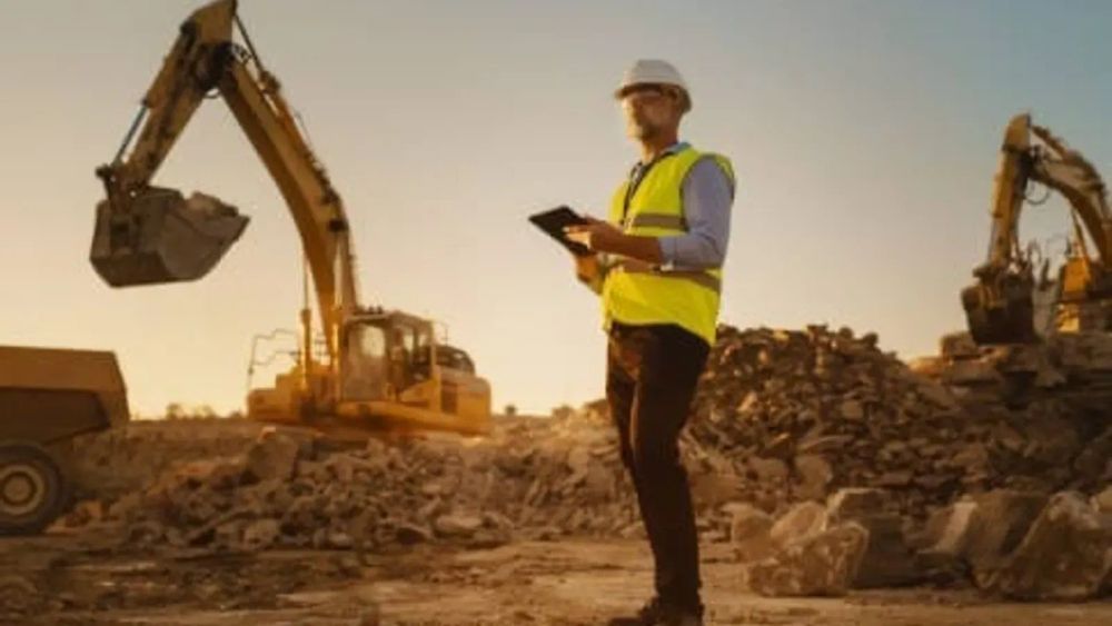 Un ingénieur en casque et gilet de sécurité examine une tablette sur un site minier avec des excavatrices, évoquant l'extraction du lithium.