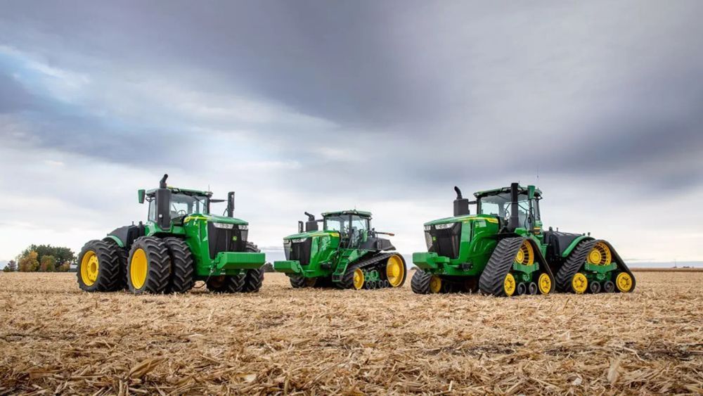 Trois tracteurs John Deere verts et jaunes dans un champ labouré sous un ciel nuageux.