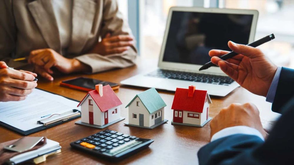 Des personnes discutent de l'investissement immobilier avec des modèles de maisons, un ordinateur portable et une calculatrice sur la table.