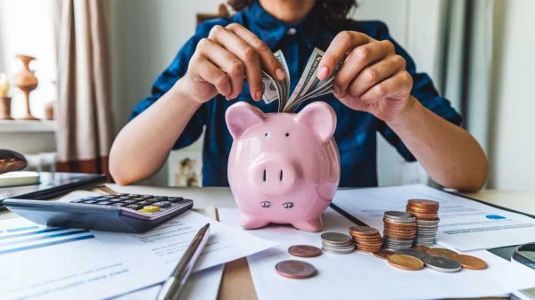 Une personne met des billets de banque dans une tirelire en forme de cochon rose, entourée de pièces de monnaie, d'une calculatrice et de documents financiers.