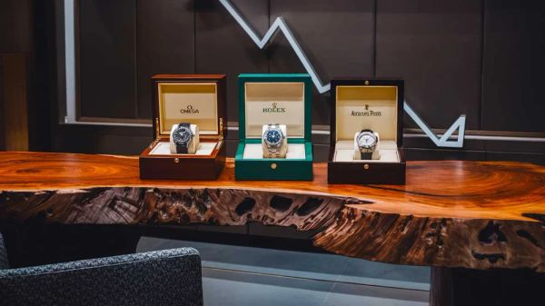 Trois montres de luxe (Omega, Rolex, Audemars Piguet) présentées dans leurs boîtiers respectifs sur une table en bois massif.