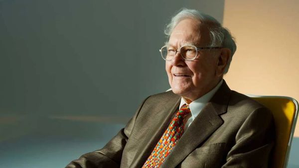 Warren Buffett souriant, portant un costume marron et une cravate colorée, assis sur une chaise jaune.
