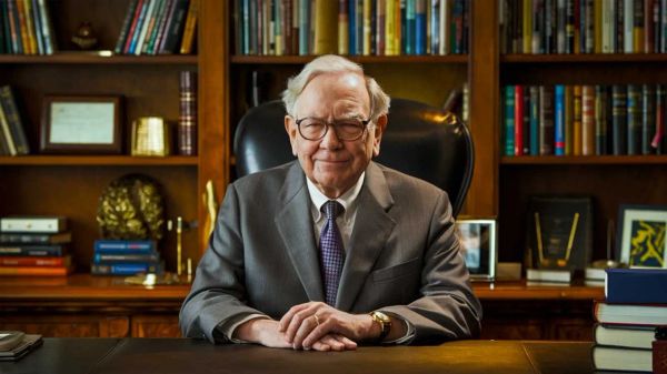Warren Buffett assis à son bureau, les mains croisées, devant une bibliothèque bien garnie.