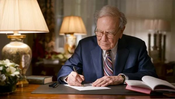Warren Buffett, assis à son bureau, rédigeant un document avec un stylo, entouré de lampes et de livres.