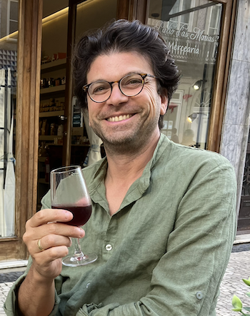 A man with dark hair and darkish olive skin grins at the camera. He is wearing a green linen shirt and holding a glass of red wine.