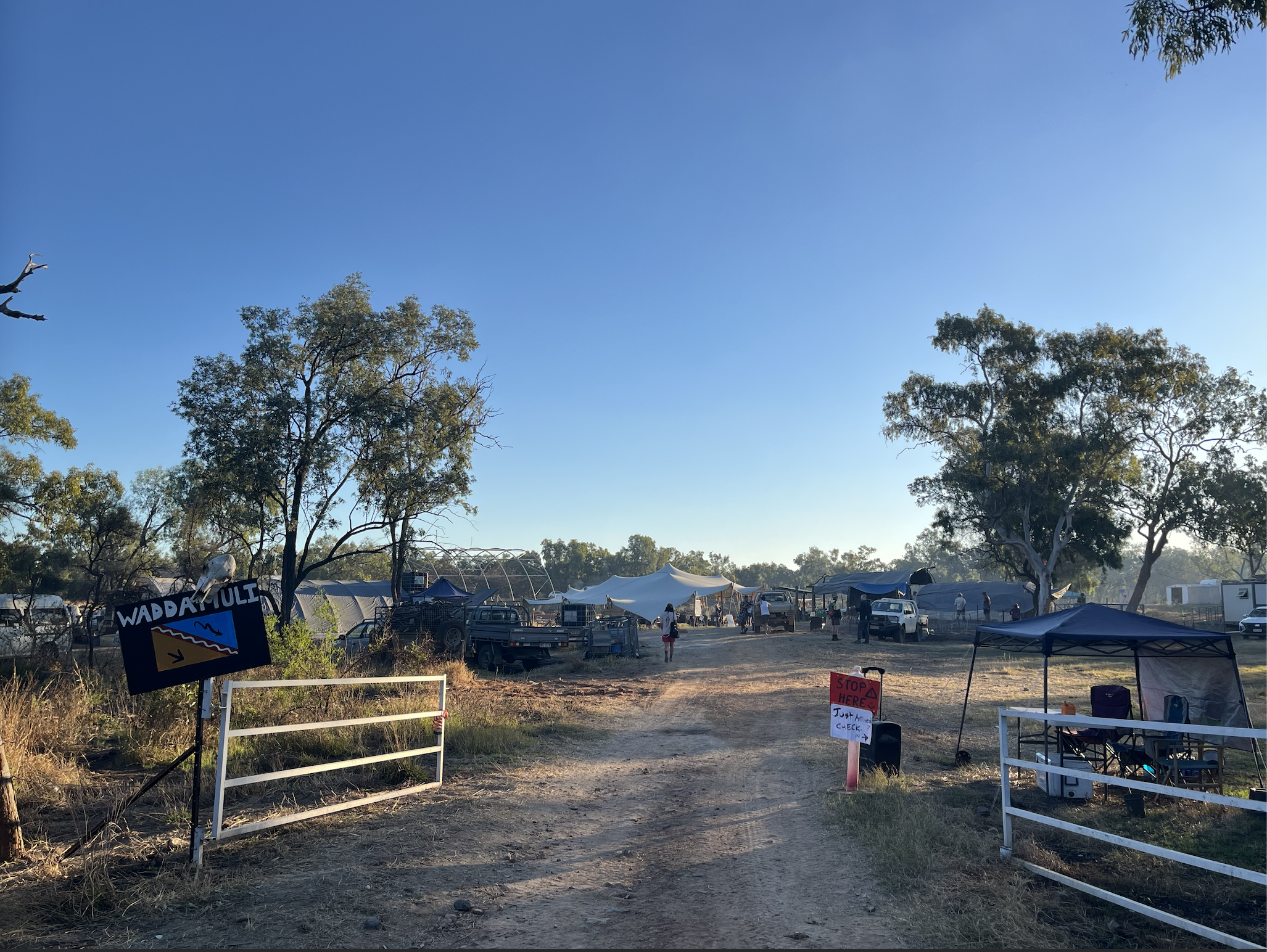 A dirt road runs between open gates. A sign at the left says "Waddamulli". Tents stand among trees under a blue sky.