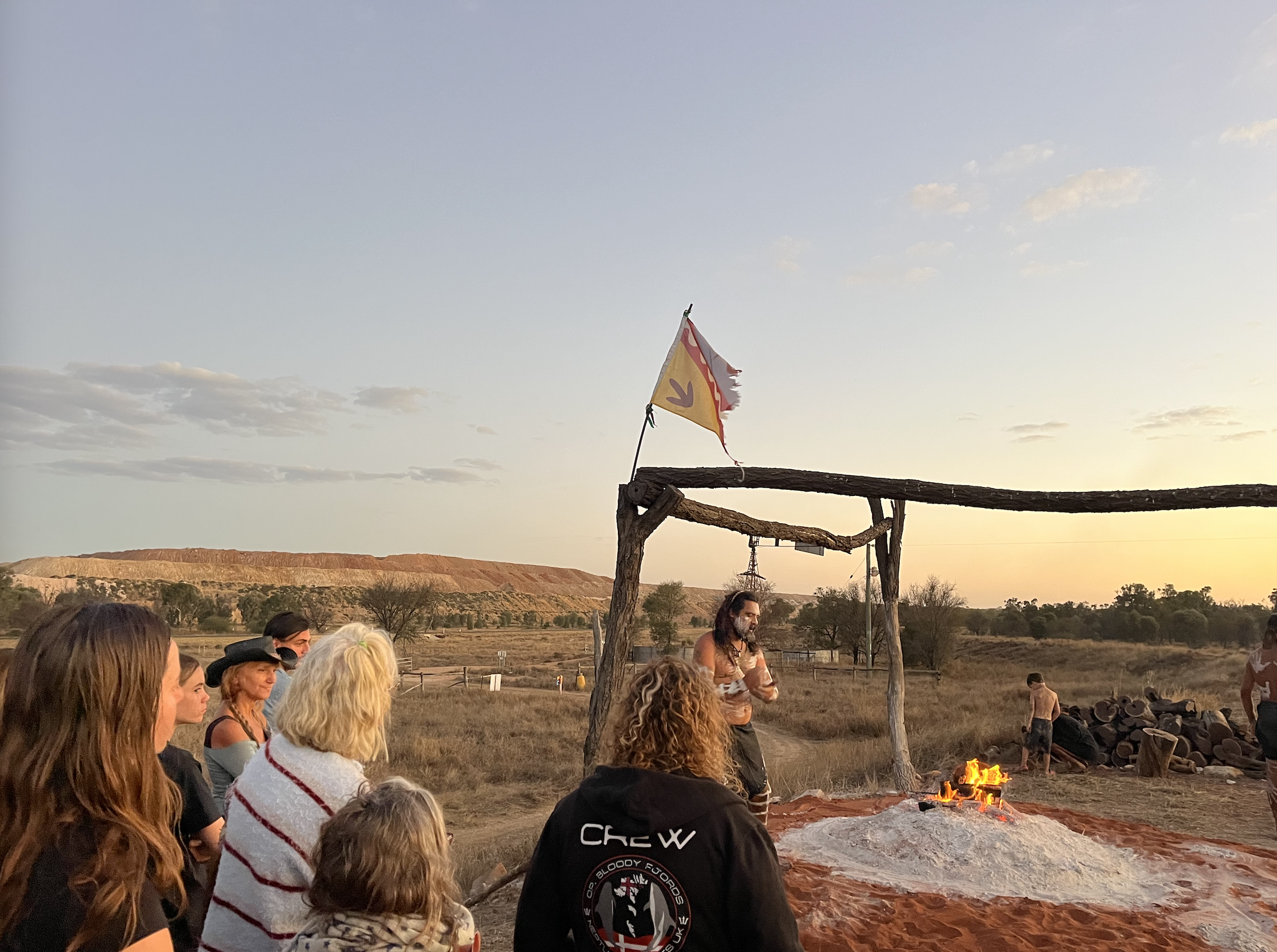 Gurridyula stands in front of the buri (fire) that is still burning. A tattered Waddananggu flag flies. The Carmichael mine looms across the road.