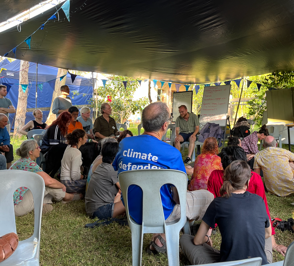 A group of people in a messy circle engaging in the "soft skill" of collaboration, under a tarpaulin on a grassy floor, with whiteboards