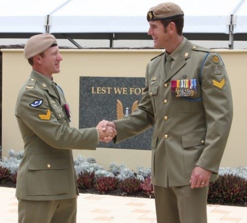 A very tall man and a much shorter man in khaki uniforms with many colourful medals, shaking hands and smiling in front of a sign reading "Lest We Forget"