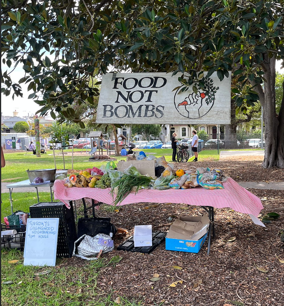 A trestle table under a Port Jackson fig tree (I think), piled high with reclaimed food. A banner hangs behind saying "Food Not Bombs"
