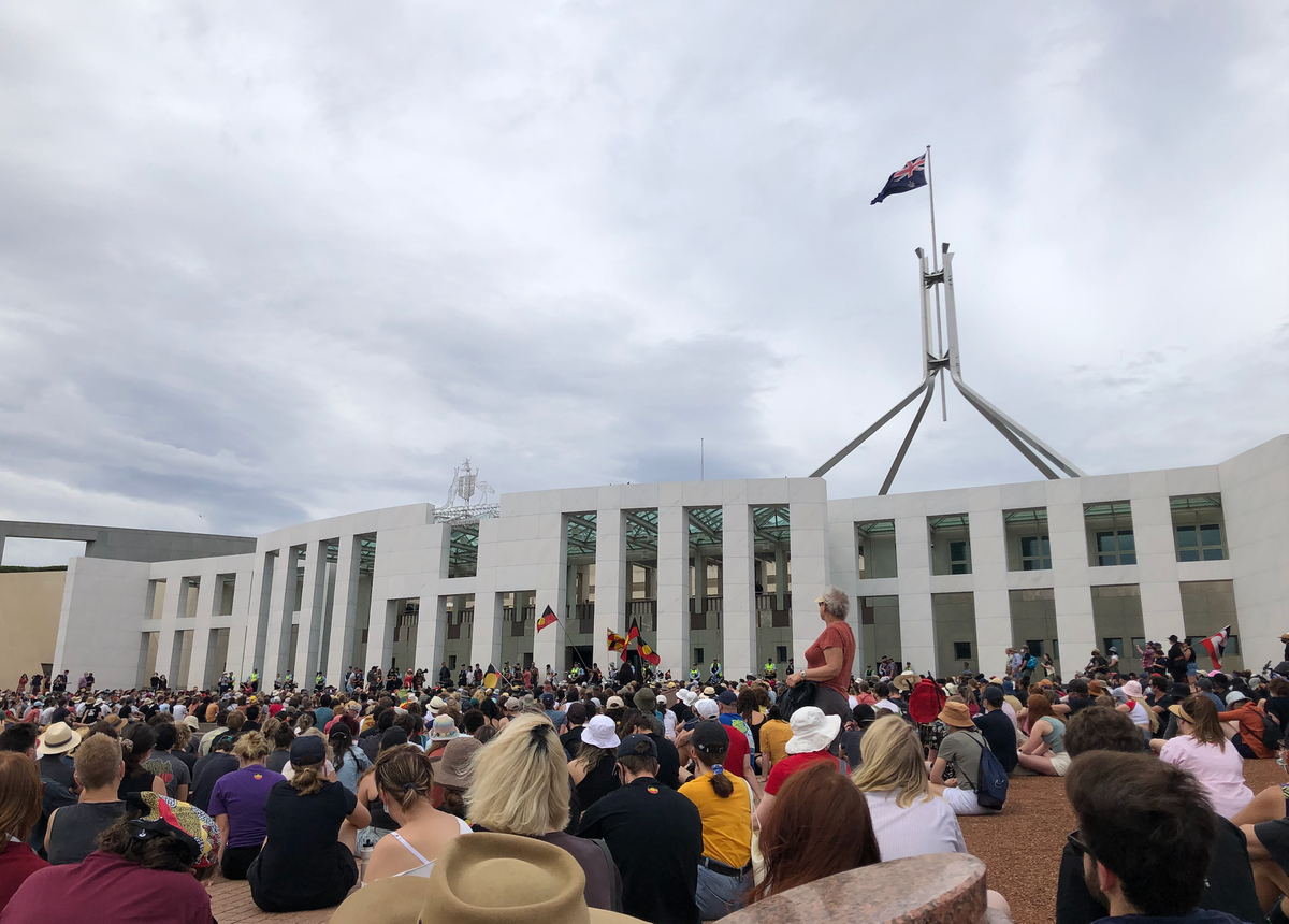 A crowd of people in bright colours sitting on the forecourt outside Parliament. By the portico, big Aboriginal flags are being waved.