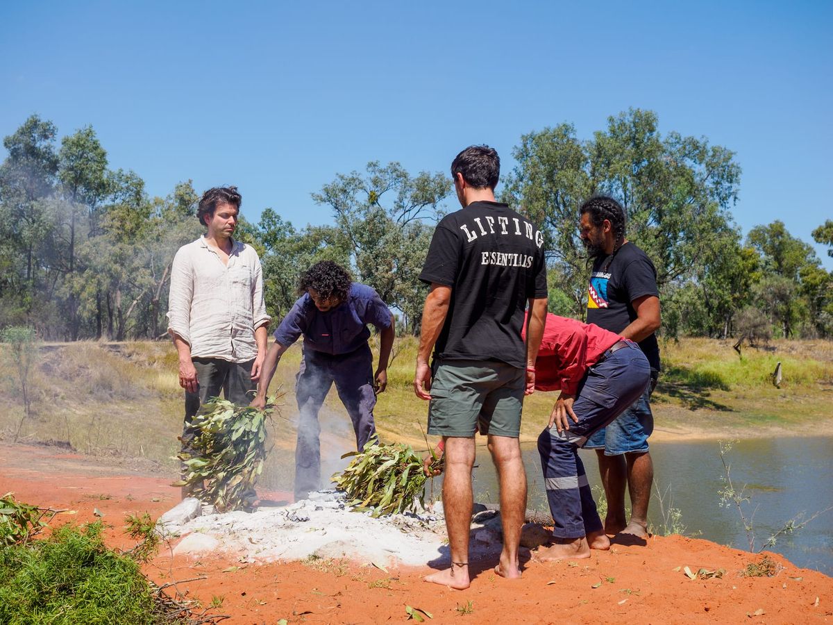 Under a blue sky, standing on red dust in front of a sacred buri (fire), I am being smoked in by Gurridyula (long hair on the right) and his brothers.