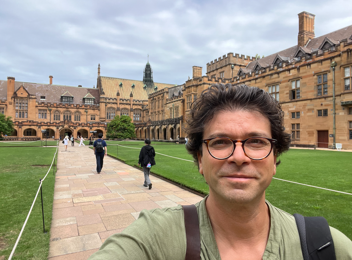 Selfie of a guy with dark hair and glasses smiling at the camera inside the Hogwartsy Quad of Sydney Uni