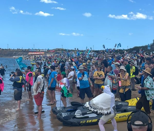 A performance activist in a seagull suit dances in front of the cops (out of shot), facing a HUGE crowd on people in colourful clothes on the beach, looking out at the water under a blue sky