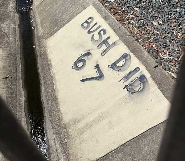 A concrete stormwater drain with "BUSH DID 6 7" graffitied on the side. A trickle of water in the bottom