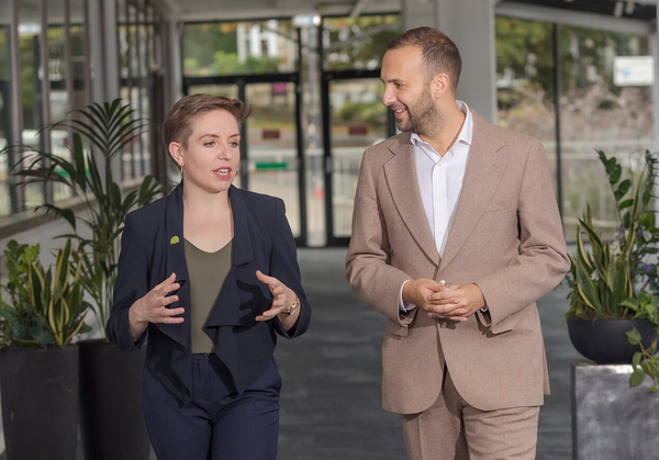 A youngish woman in a dark blue suit gesturing and speaking, with a youngish man in a tan suit looking at her admiringly