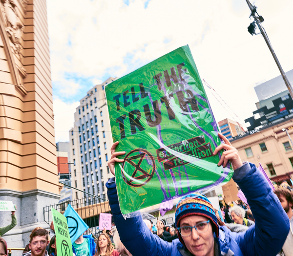 An XR protester wearing a beanie and puffer jacket holds a placard reading "Tell the truth"