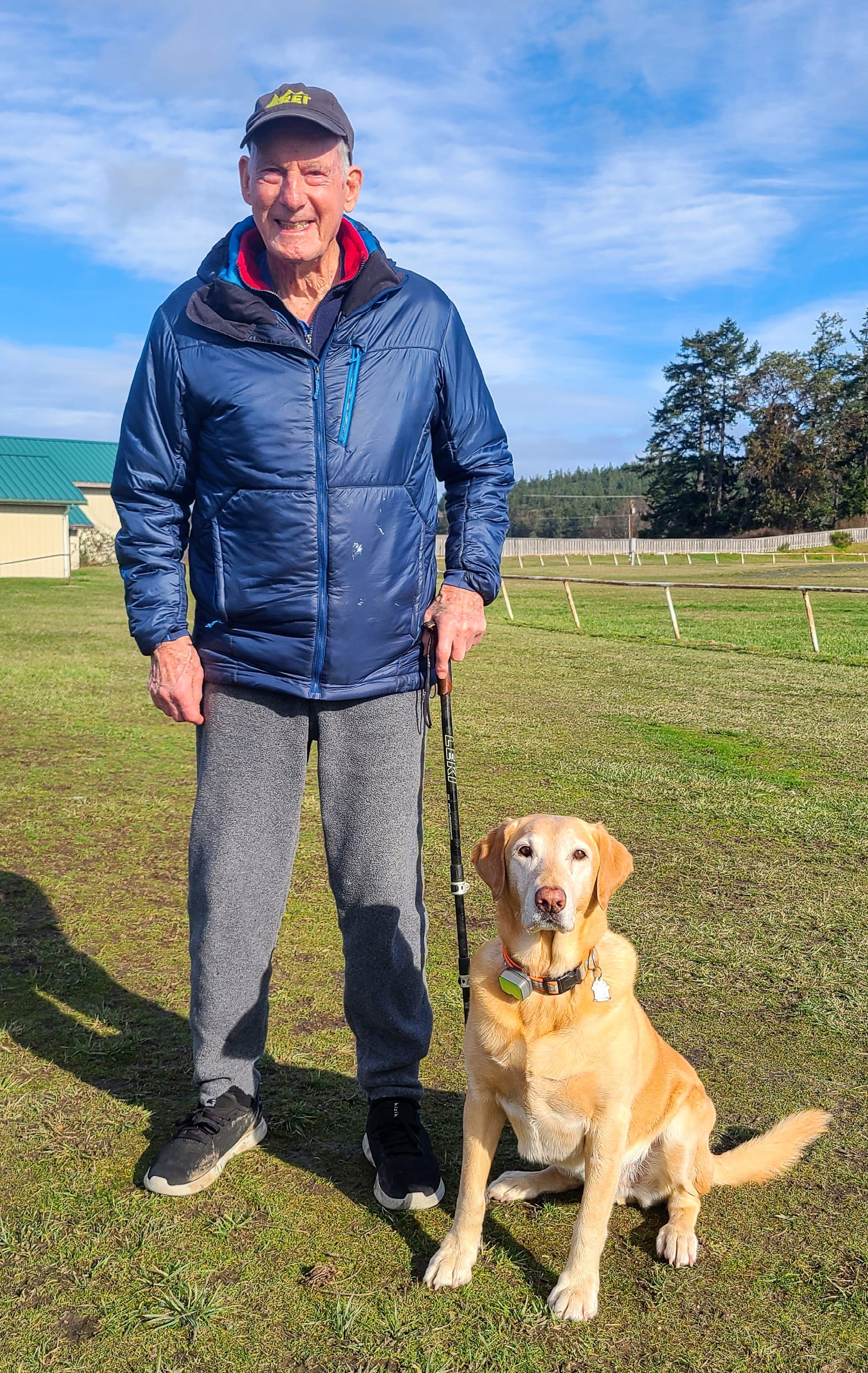 Jim in a blue coat with gray pants and his yellow lab in the grass at the fairgrounds