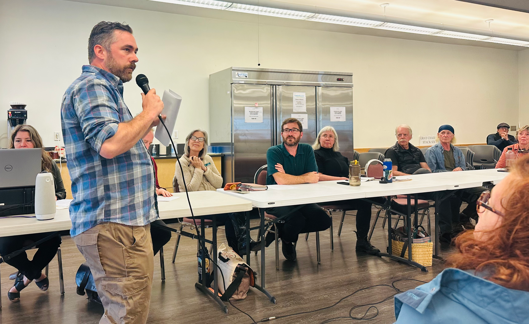 Devon Buckham speaking into a microphone standing to the left as seated listeners look on.