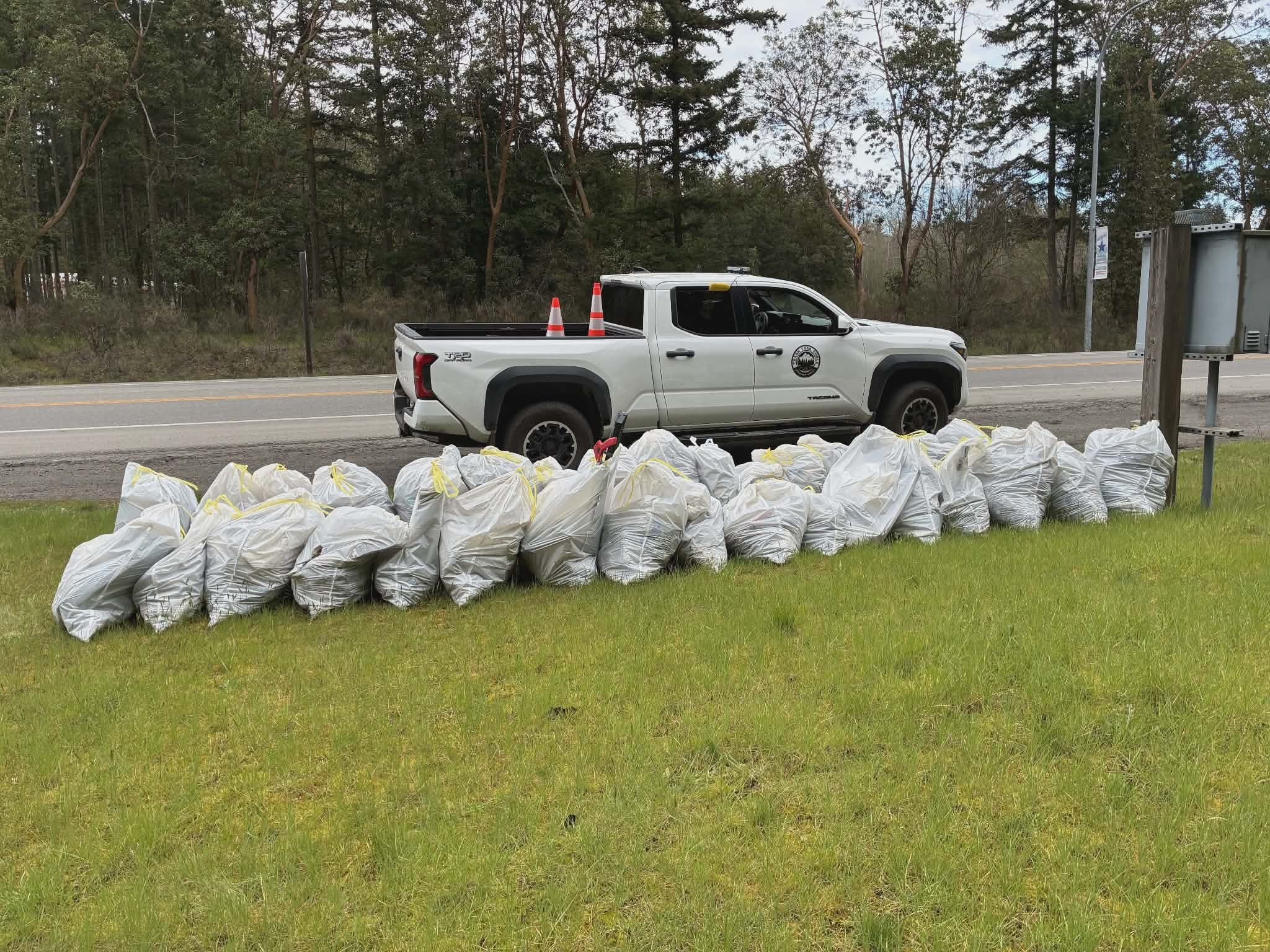A pile of white litter bags in front of a white truck along side of the highway. 