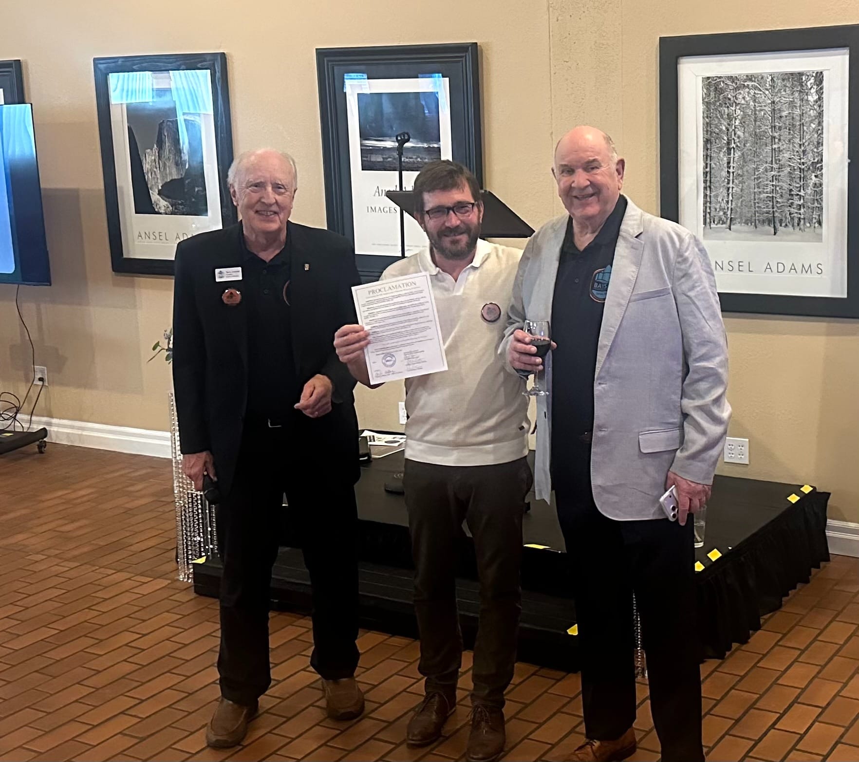 Terry Umbreit, Greg Brotherton, and Gary Keister standing in front of a small stage with a mic a proclamation and a glass of wine for a toast.