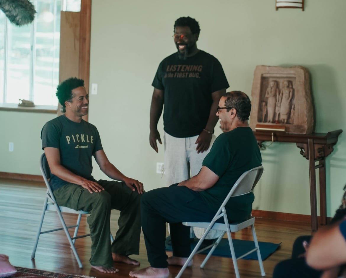 Two men facing each other in folding chairs with one standing and looking on. 