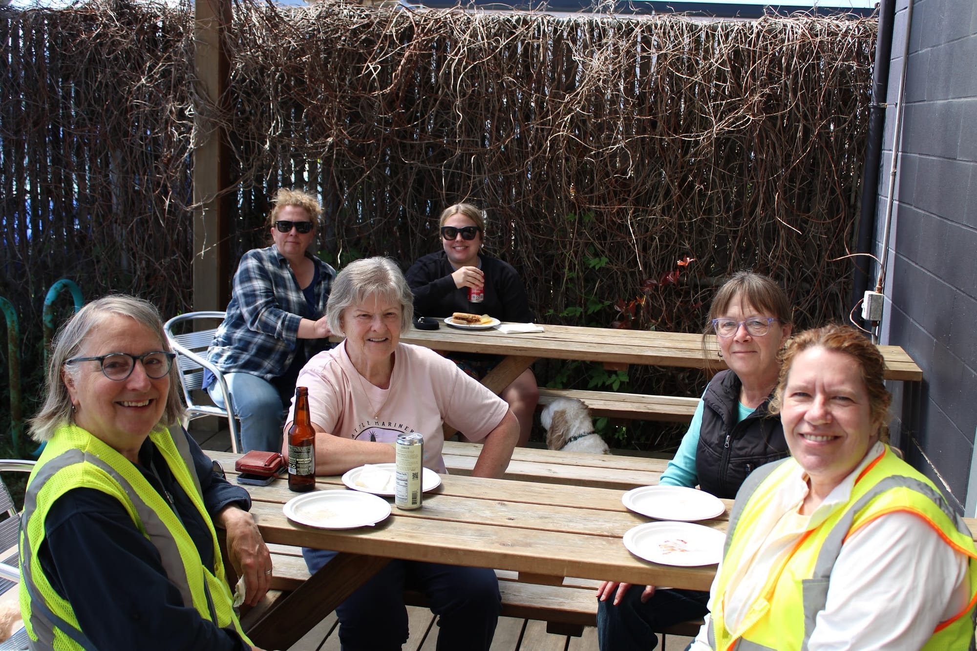Volunteers in reflective vests in the foreground with others seated behind at picnic tables in front of empty paper plates and beverages. 