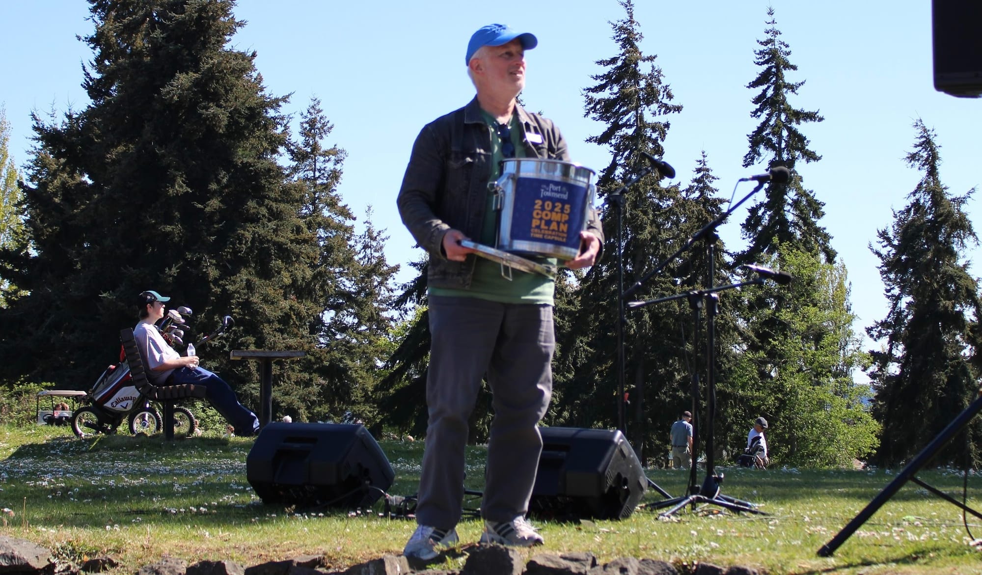 Deputy Mayor Owen Rowe holding a silver time capsule with fir trees and onlookers in the background. 