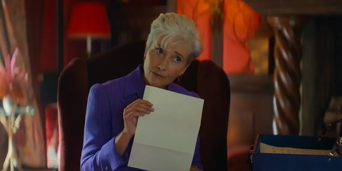 Emma Thompson is holding a piece of paper wearing a purple jacket sitting in a high-backed chair.