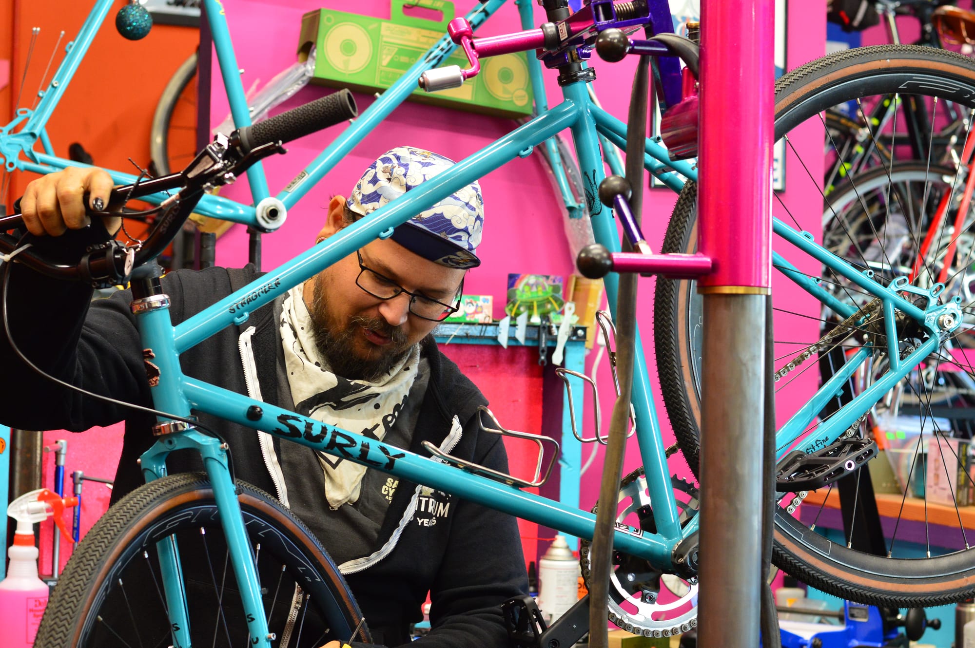 Gabe Quintanilla works on a blue bike on a bike stand with pink wall in the background. 