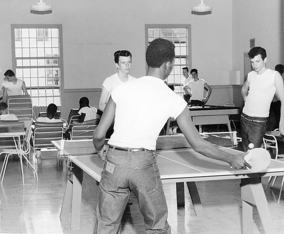 A black and white photo of boys in jeans and white t-shirts playing table tennis and lounging in the background.
