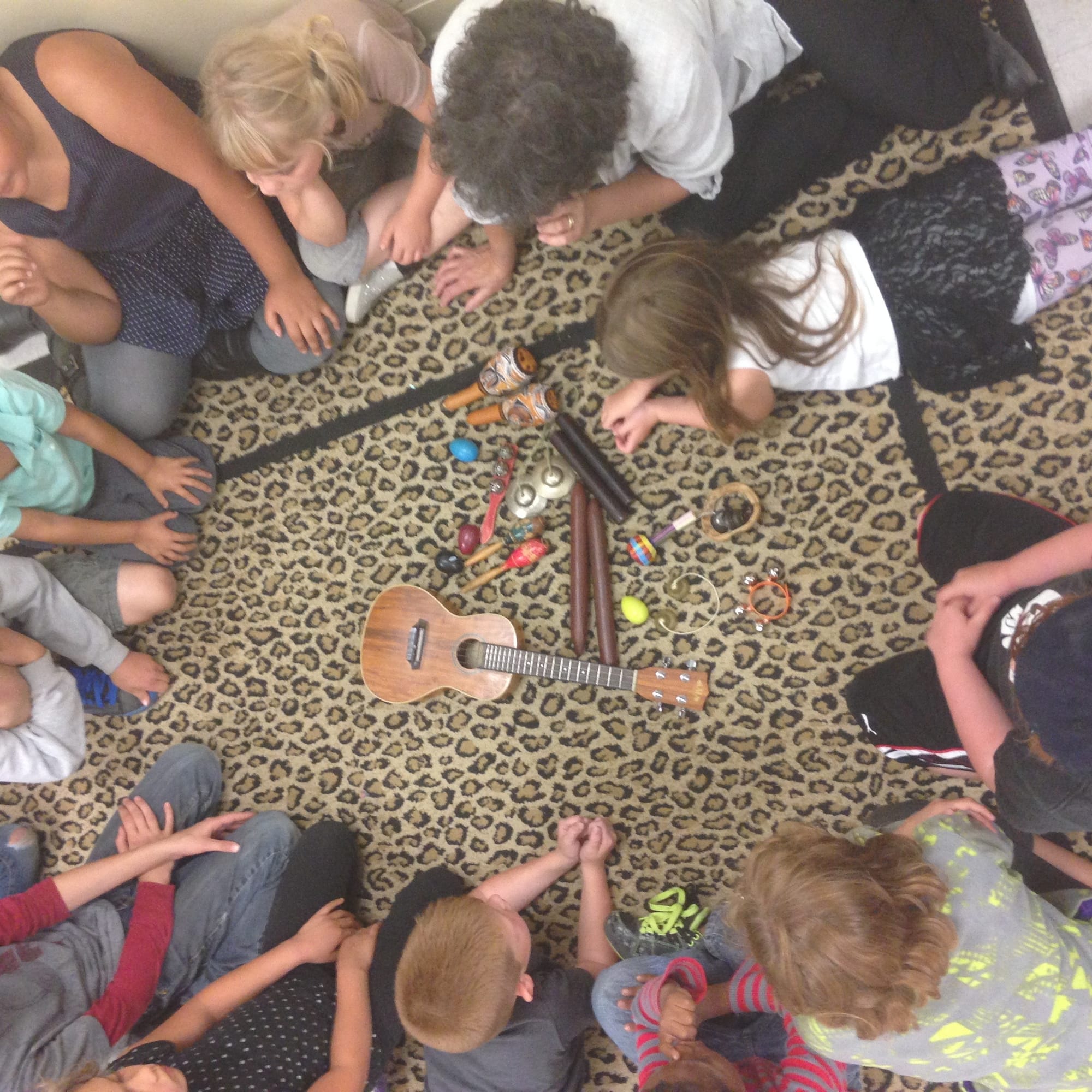 An overhead picture of kids in a circle on a leopard print rug looking at percussion instruments and a ukelele.