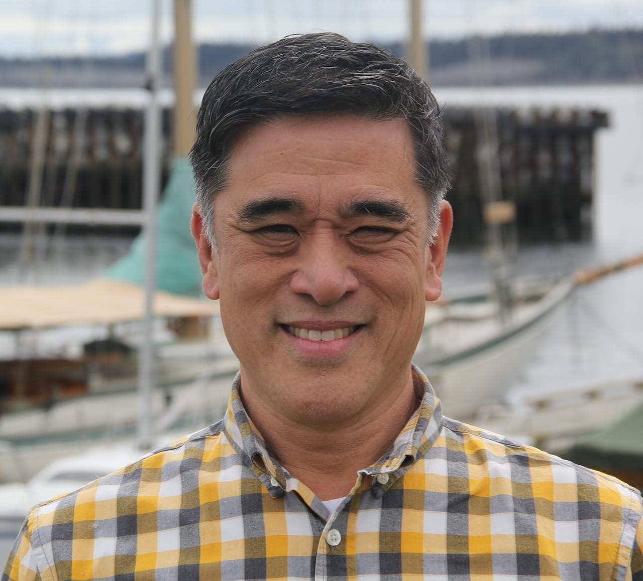 Dave Nakagawara in a yellow and black plaid shirt standing on the beach in front of the Port Townsend Marina breakwater.